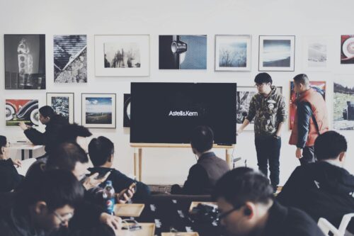 A group of adults attending a casual business presentation in a modern art space, featuring a TV display and framed art.