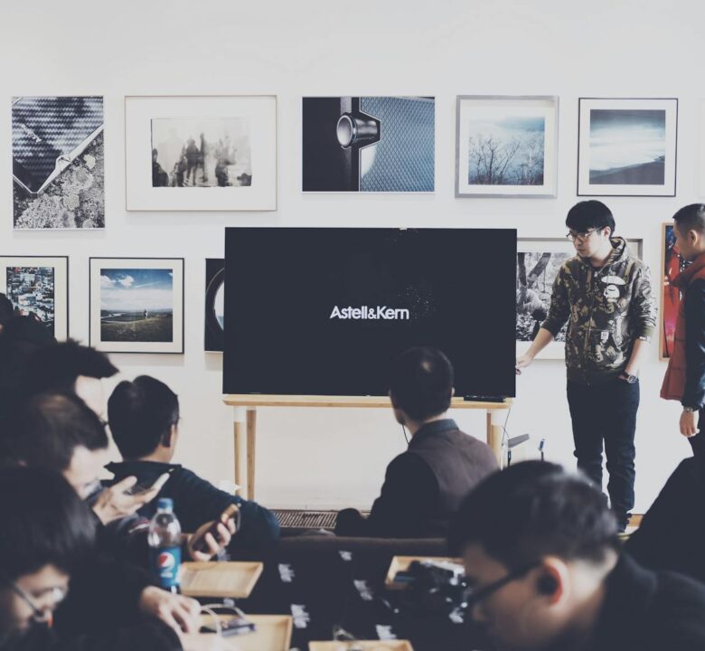 A group of adults attending a casual business presentation in a modern art space, featuring a TV display and framed art.